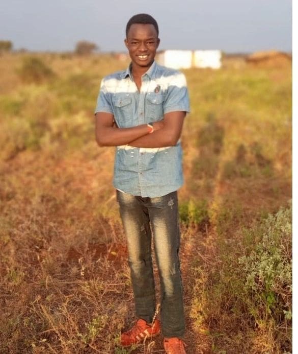A man standing in an open field with dry grass, smiling and wearing casual clothes.
