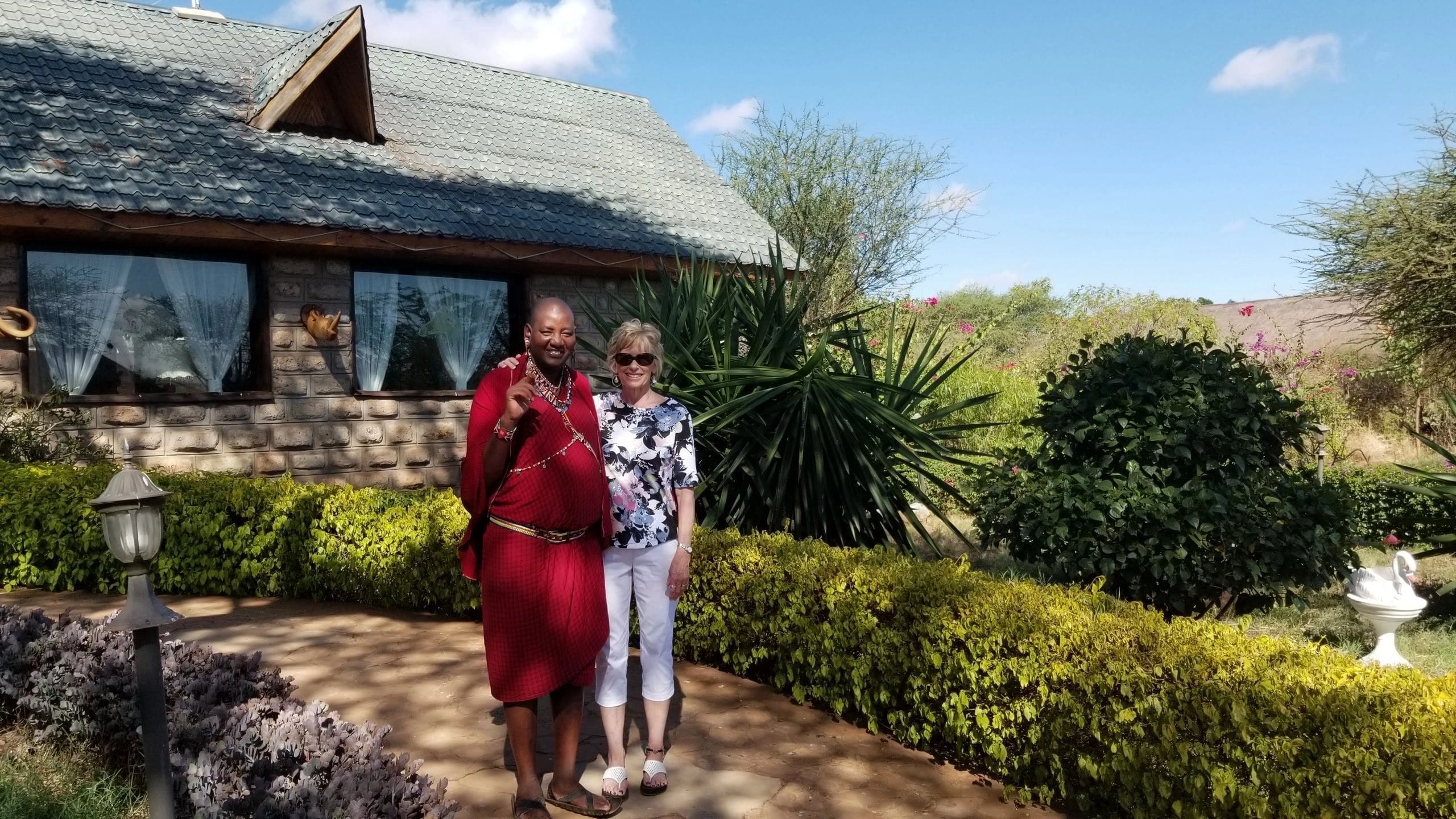Two women enjoying a sunny day in a lush garden outside a rustic house.