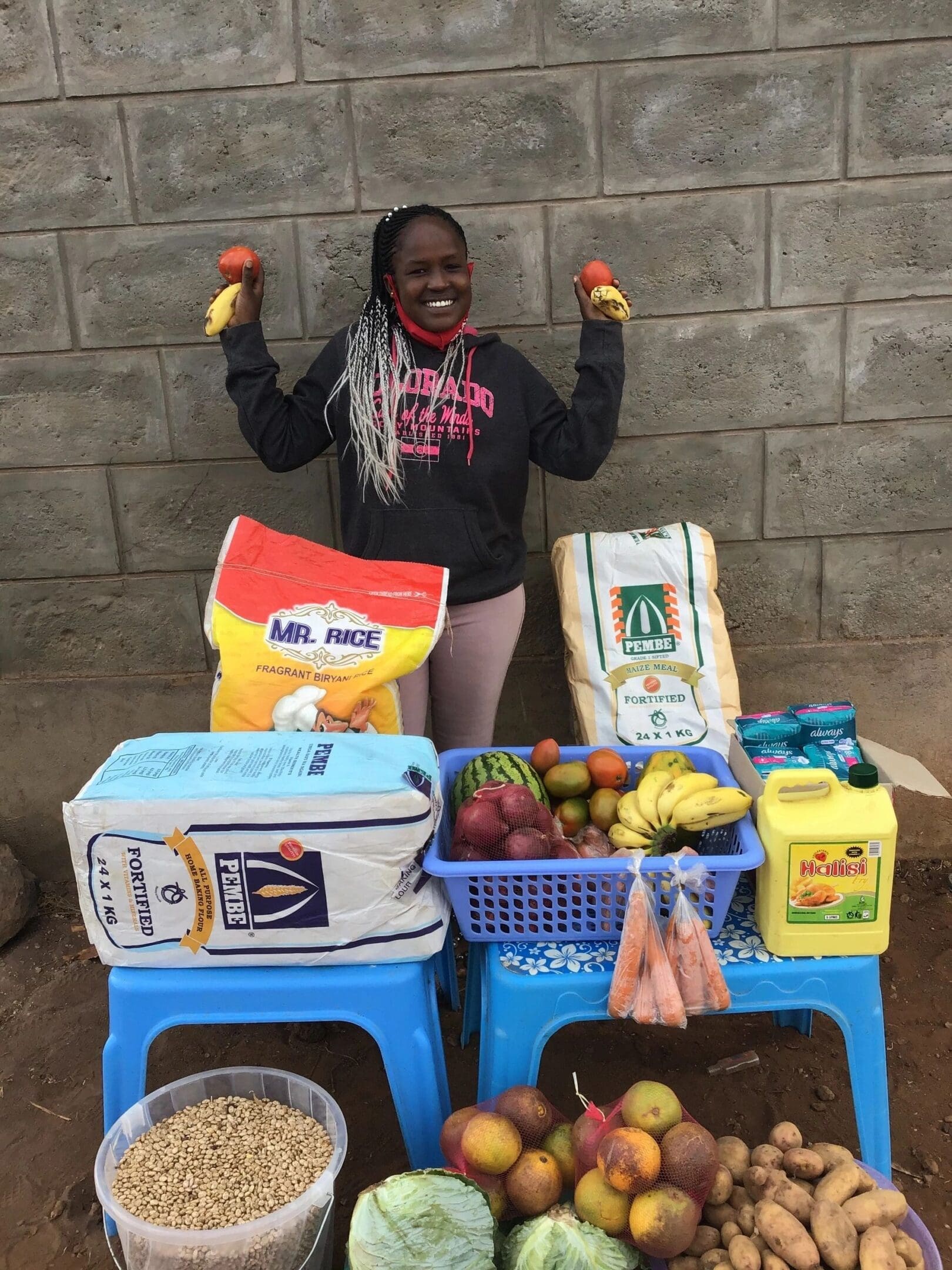 Smiling girl proudly displays groceries on crates against a brick wall.