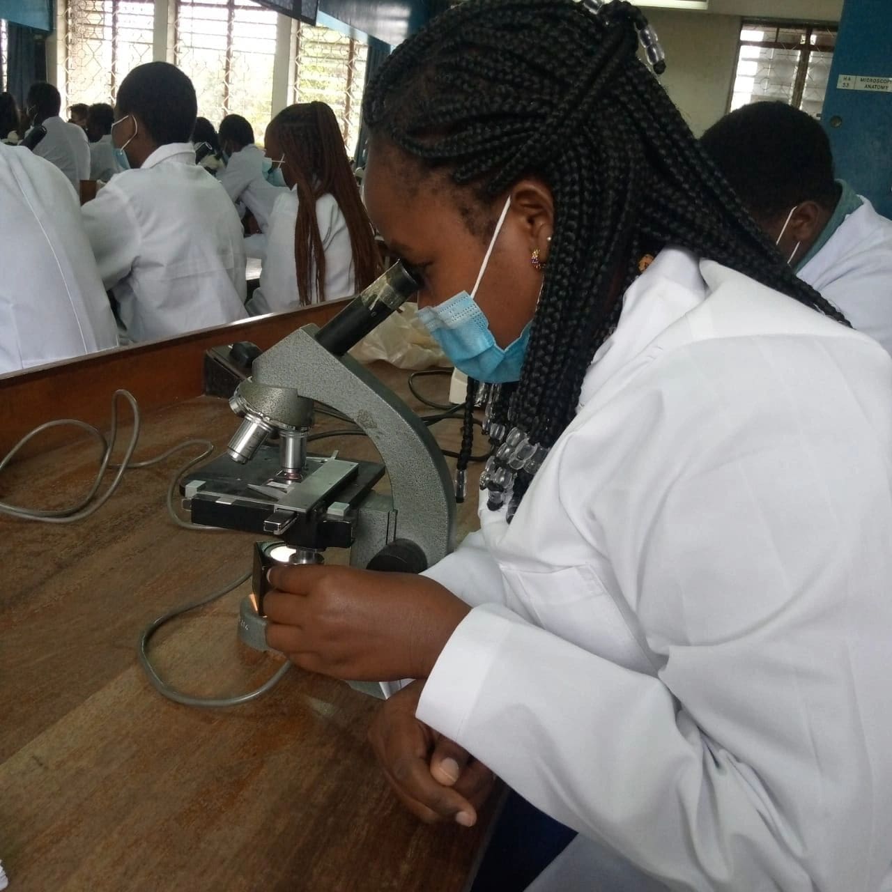 A student in a lab coat using a microscope in a classroom.