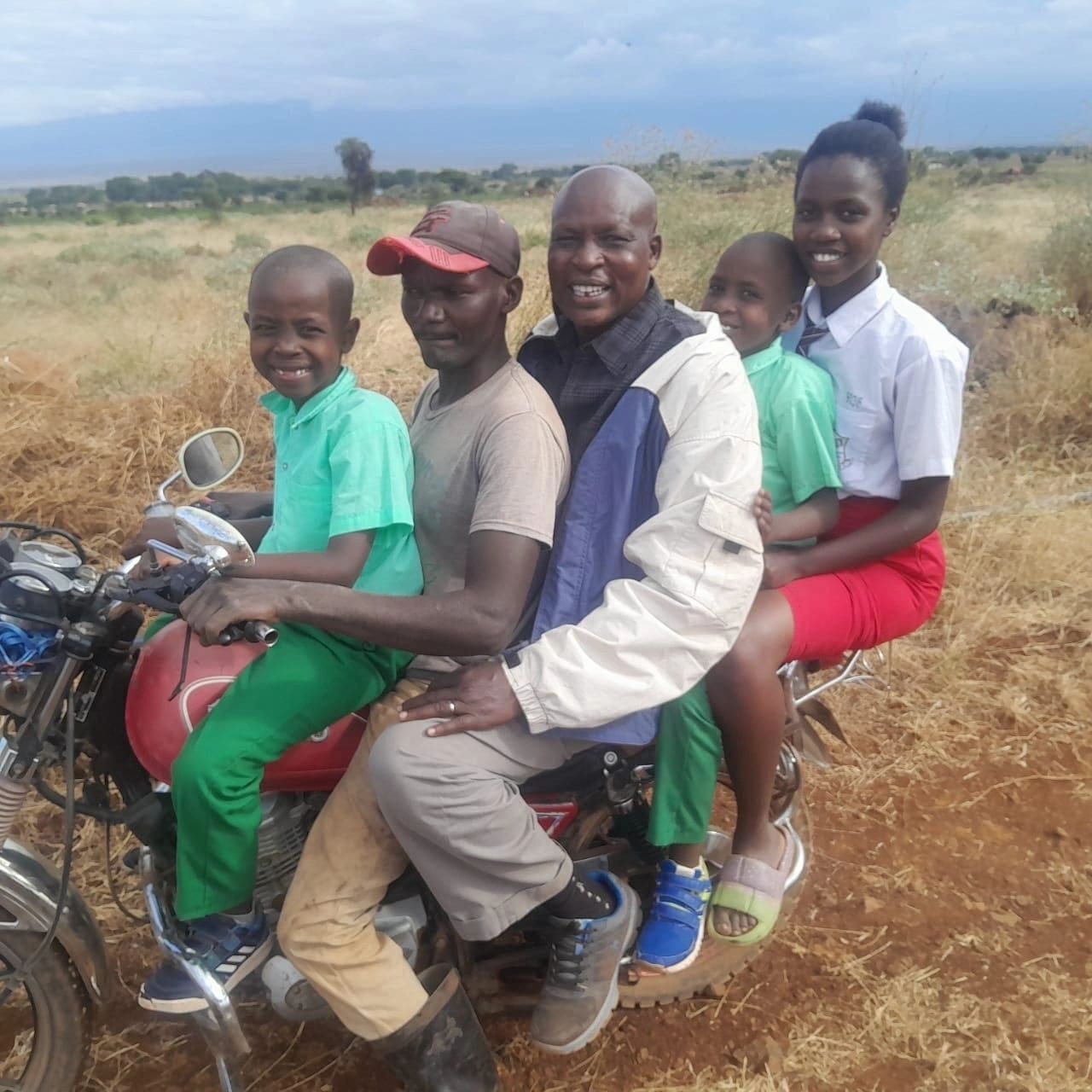 Five people riding a motorcycle on a dirt road in a rural area.