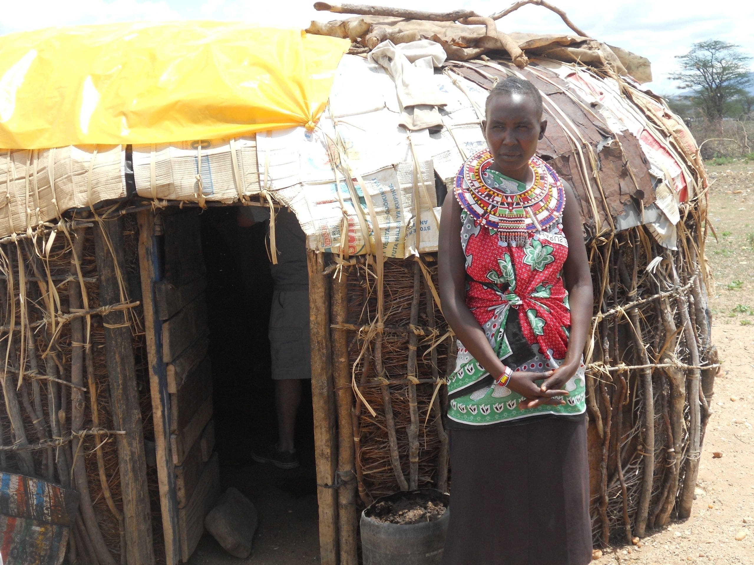 Woman standing outside a traditional hut with a thatched roof.