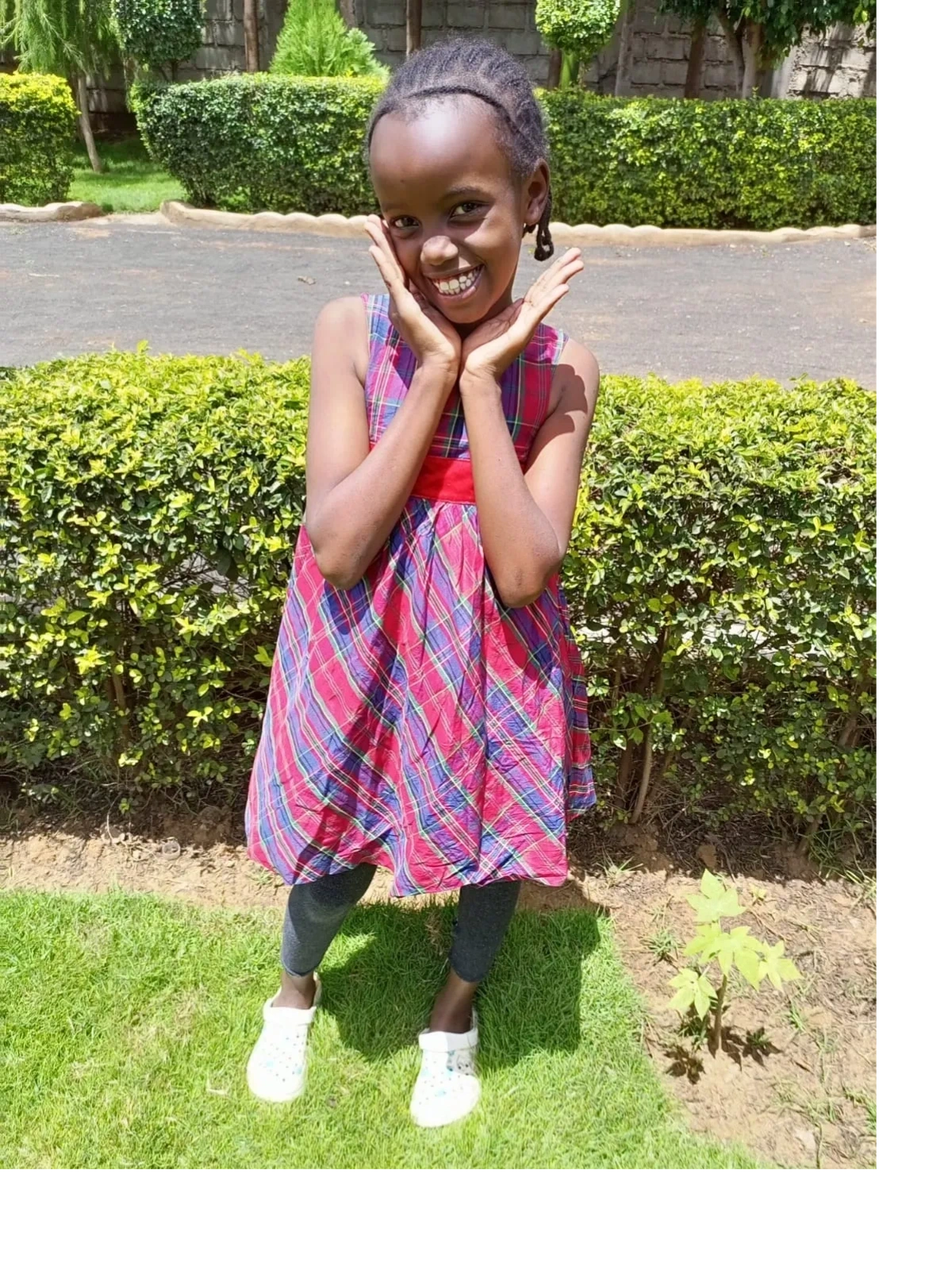 A young girl in a red plaid dress poses happily outdoors.