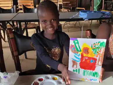 Smiling girl proudly shows her colorful painting at a table.
