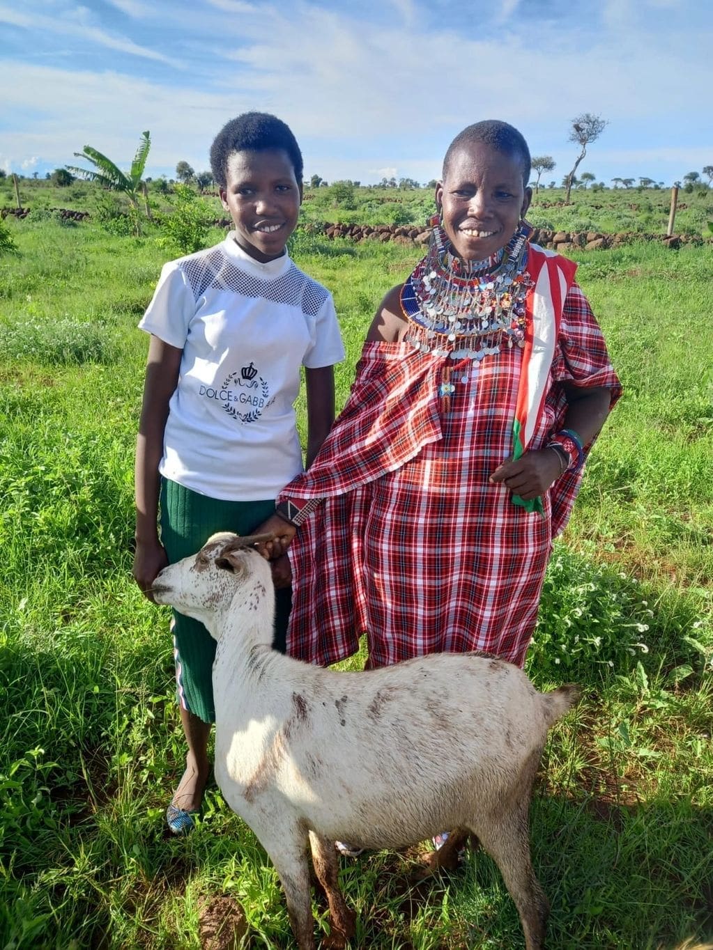 Two women smiling outdoors with a goat in a green field.