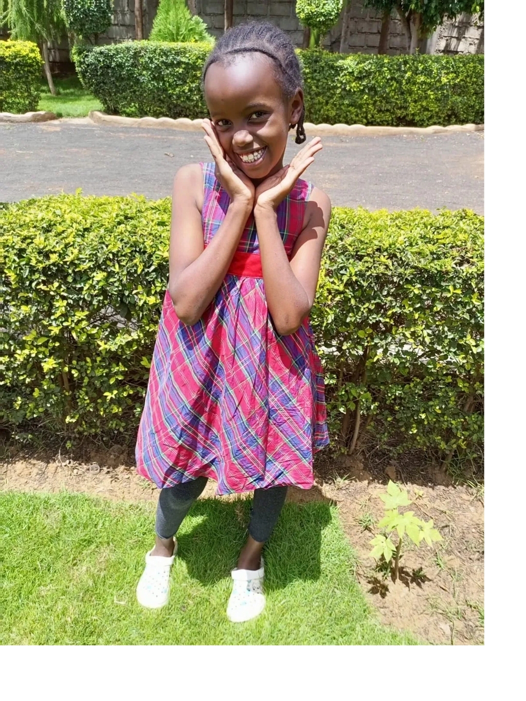 A smiling girl in a red and blue plaid dress posing outdoors on grass.
