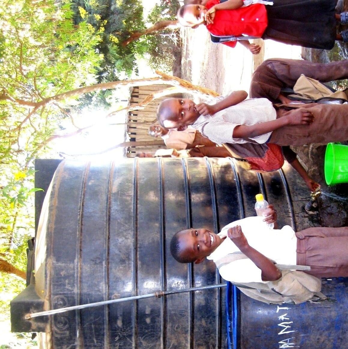Children filling containers from a large water tank outdoors.