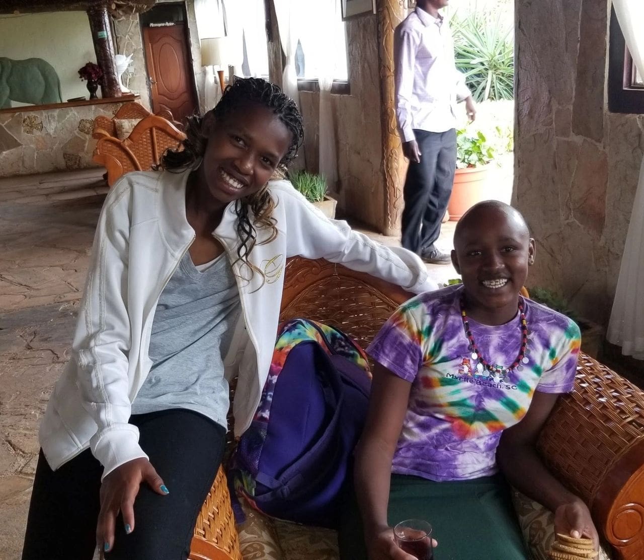 Two women smiling together indoors, one sitting and one standing.