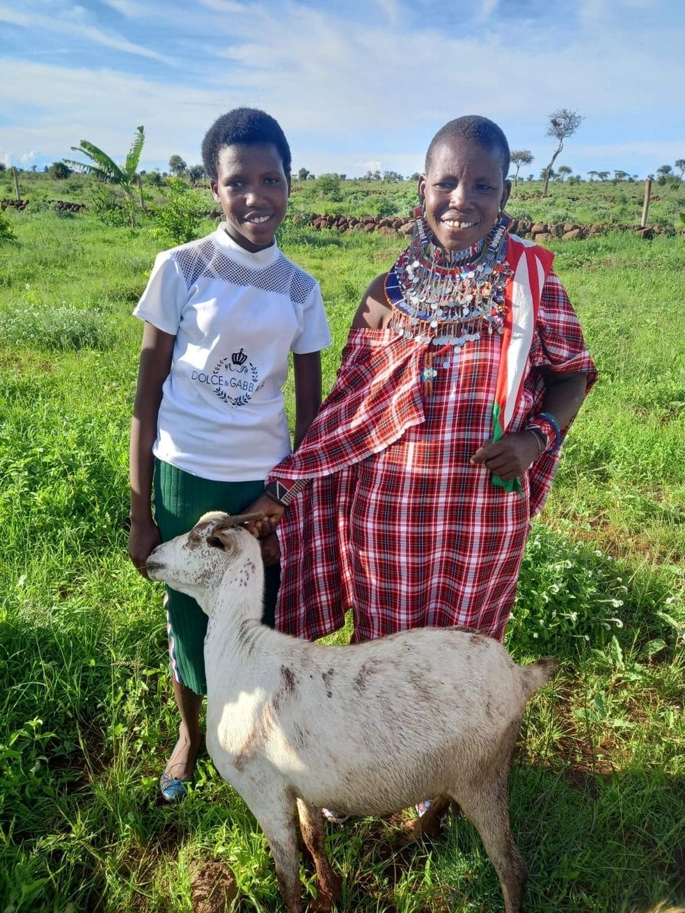 Two women standing outdoors with a goat in a green field.