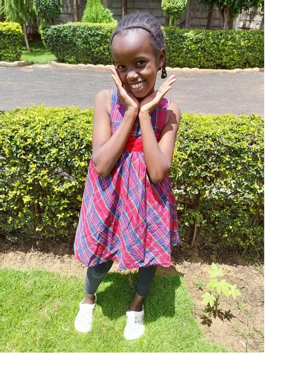 A smiling girl in a red plaid dress poses outdoors on grass.