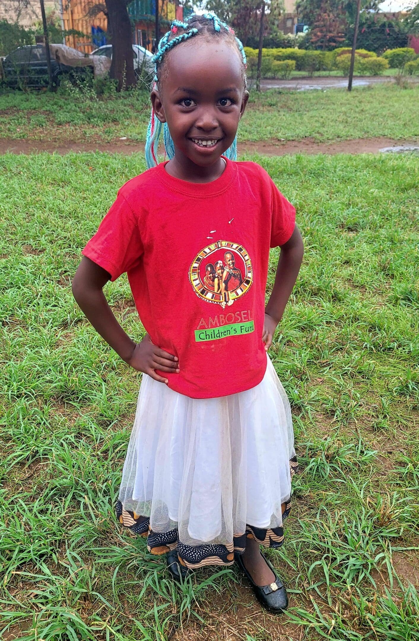 A smiling girl in a red t-shirt and white skirt stands on grass.