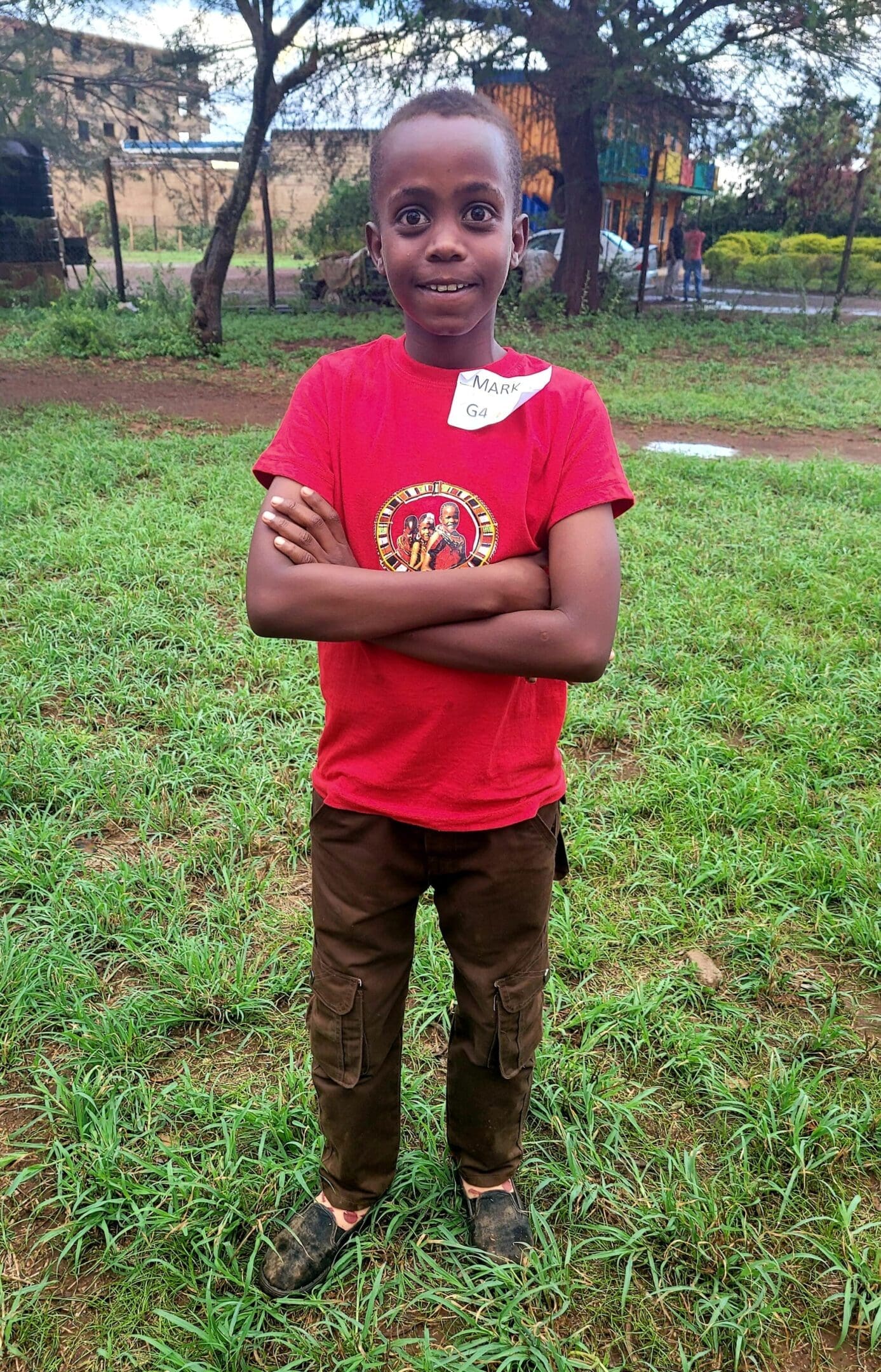 A young boy in a red shirt standing on grass with arms crossed.